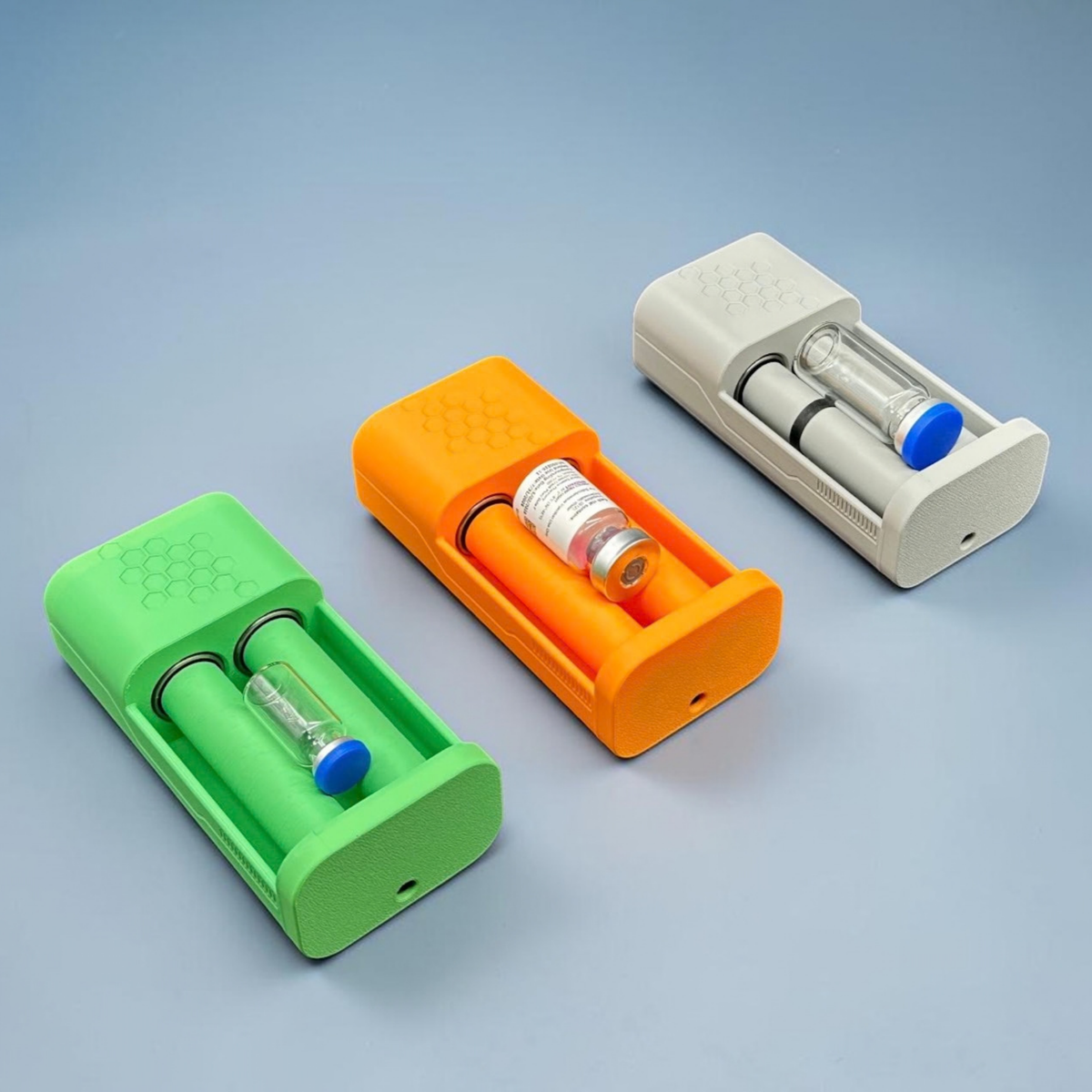 Three open, motorized, vial roller holders in different colors (matte gray, matte mandarin orange, and matte grass green). Each device shows a vial nestled between two rollers. The vials are three different sizes. The devices are lined up in a row but oriented diagonally, pointing down towards the right corner of the frame.