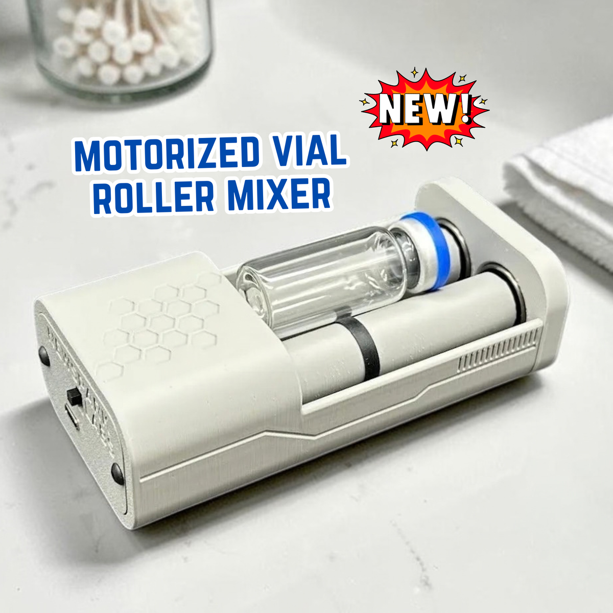 An open, gray motorized vial roller mixer device shown on a light surface in a bathroom. Blue text reads 'MOTORIZED VIAL ROLLER MIXER' and a red starburst says 'NEW!'. A small vial with a blue cap lays in the device between two rollers. A decorative honeycomb motif is etched into the top and there are decorative ridges on the side. 